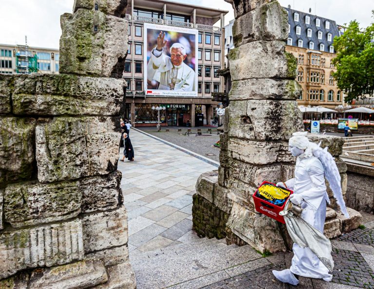 Engel mit Bierkasten vor dem Papst. Eine lebende Statue schreitet mit einem Bierkasten, den sie als als Podest verwenden wird, durch das römische Tor vor dem Kölner Dom. Im Hintergrund winkt in Überlebensgröße Papst Benedikt VI von einem Plakat / © Foto: Georg Berg