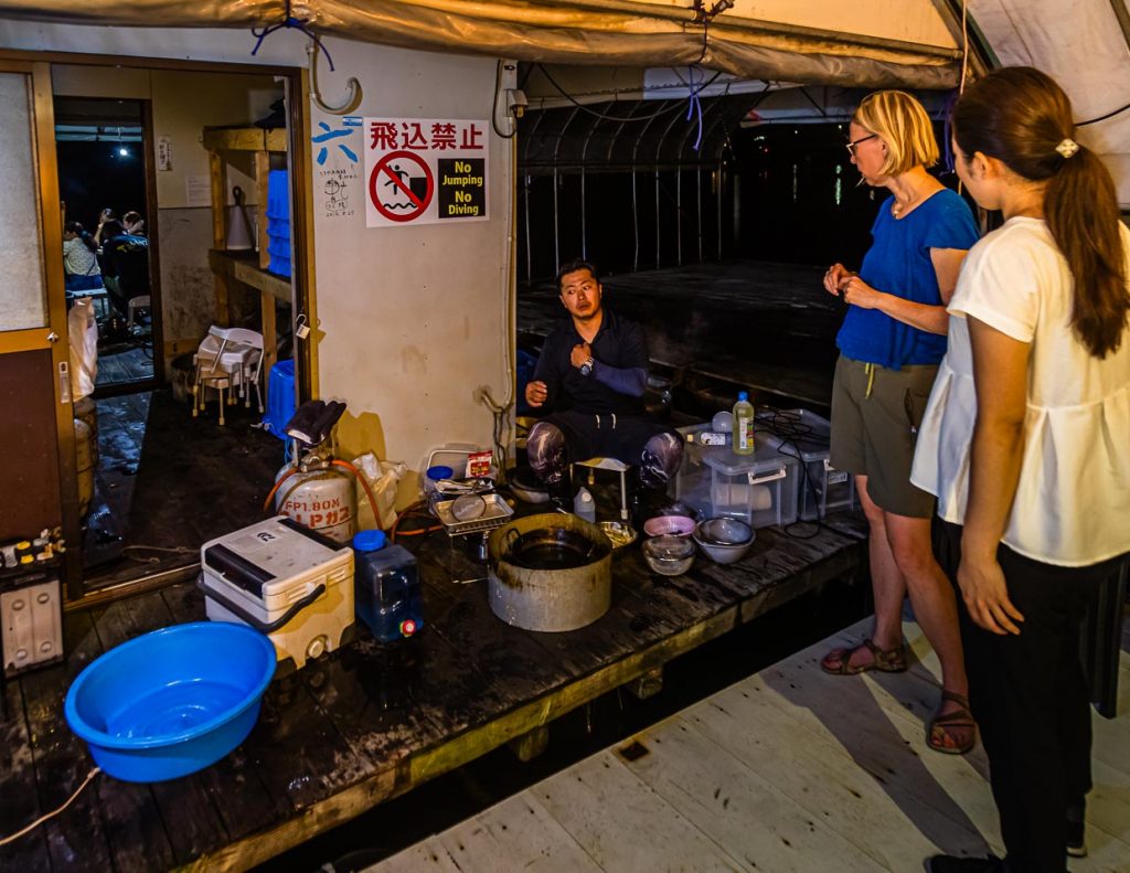 Fischer, Koch und Entertainer. Mit simpelstem Equipment begibt sich Ikuya Yamamoto an die Zubereitung des Fangs. Geschickt werden die Fische geputzt und die Krebse kommen in den Kochtopf. Gäste stellen neugierig Fragen und werden mit einem kühlen Getränk versorgt / © Foto: Georg Berg