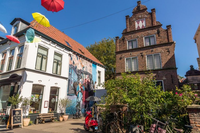 Wandmalerei in der Altstadt von Deventer. Motiv anlässlich des Hansejahres, das die neun holändischen Hansestädte 2023 feiern. Die Wandmalerei hat einen Bezug zur Vergangenheit und Gegenwart. Hier in Deventer: Marktfrau mit Sohn verkauft Deventer Honigkuchen und Stockfisch, wichtige Waren im Hansehandel von Deventer. Der Junge trägt einen Fan-Schal des Fußball-Clubs Deventer Eagles / © Foto: Georg Berg
