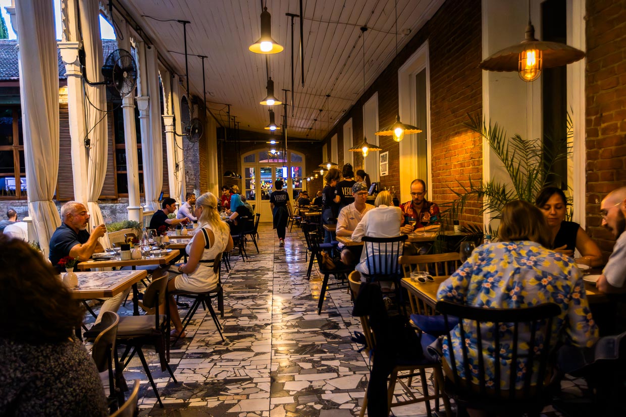 Gäste sitzen im Außenbereich der Kikodze Coffee & Cocktail Bar. Das Gebäude ist beleuchtet. Ein Bar-Schild leuchtet. Die Szene spielt sich in Vardisubani, Tbilisi, Georgien ab / © Foto: Georg Berg