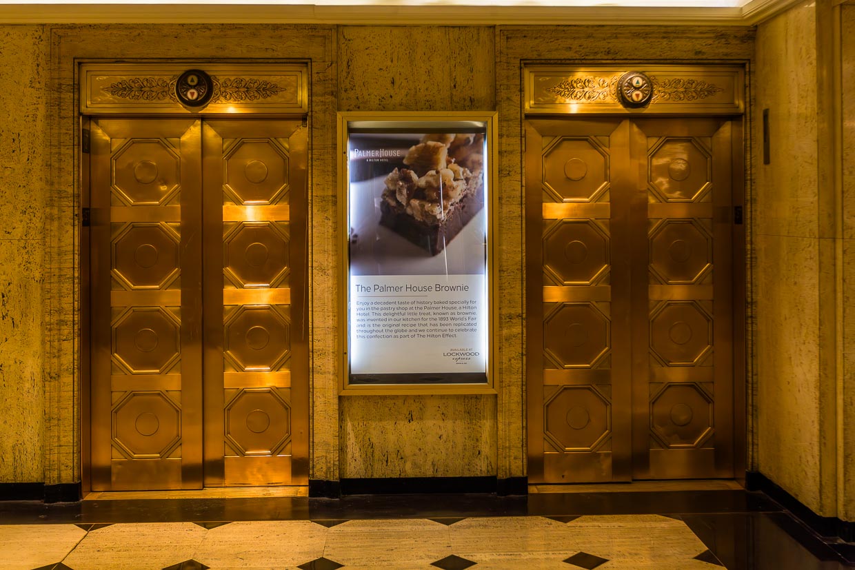 Goldene Aufzüge im Palmer House, gebaut 1873. Das Palmer House war das erste Hotel in Chicago mit Aufzügen. Hier beleuchtete Hinweistafel mit Werbung für das berühmte Gebäck, den von Bertha Palmer erfundenen Brownie / © Foto: Georg Berg