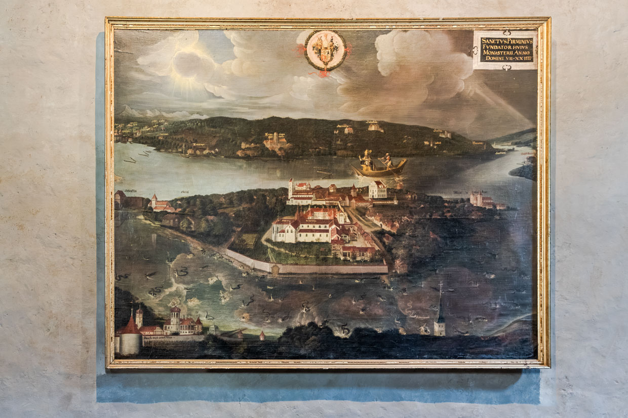 Das Tafelgemälde  von 1624 im Münster St. Maria und Markus veranschaulicht die Gründungslegende der Insel Reichenau und zeigt die Ankunft des Heiligen Pirmin. Die Bildmitte nimmt die ummauerte Klosteranlage mit der im 19. Jh. abgerissenen Abtspfalz und der Pfarrkirche St. Johann ein. Direkt am Hafen steht das „Hohe Haus“, am linken Bildrand sind die Ruine Schopflen und St. Georg in Oberzell, am rechten Bildrand St. Peter und Paul in Niederzell zu erkennen / © Foto: Georg Berg