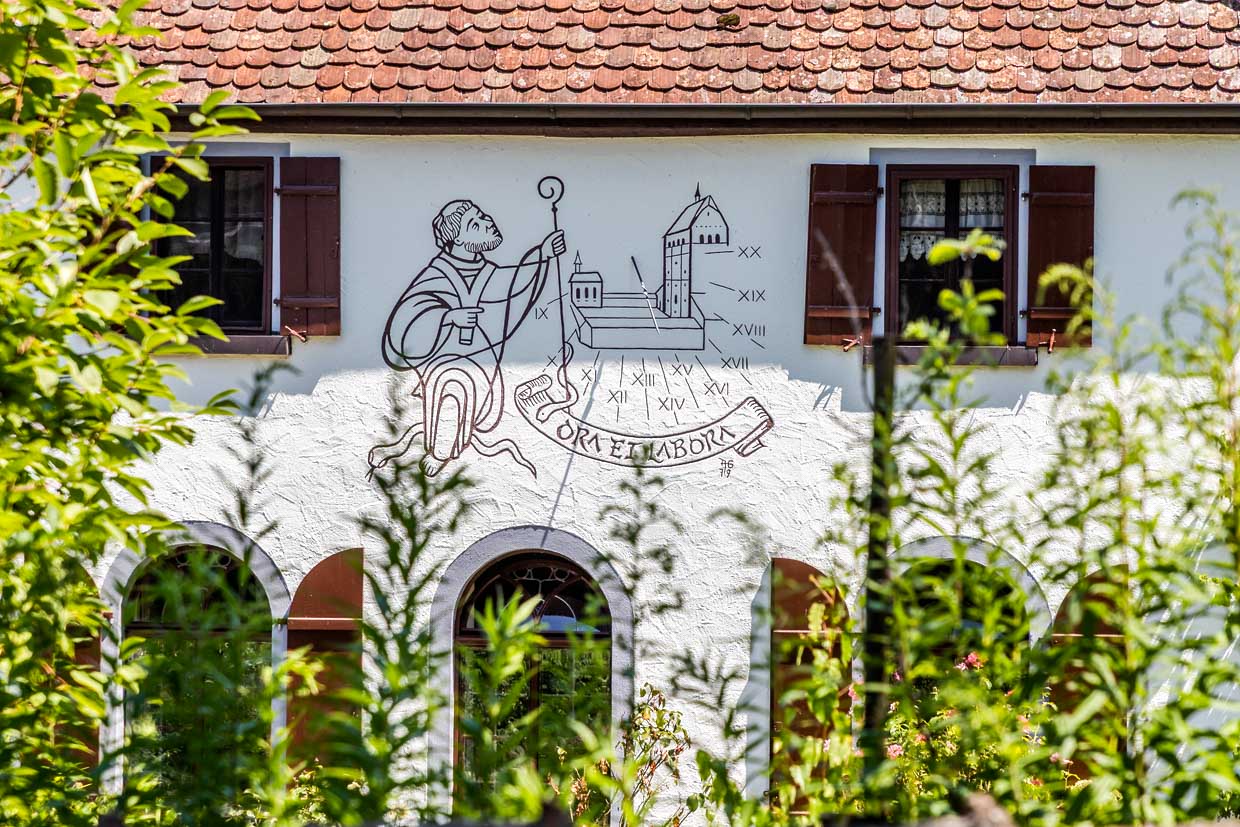 Hausfassade nahe dem Münster St. Maria und Markus in Mittelzell mit der Darstellung eines Benediktinermönches und dem Ordensspruch  Ora et Labora. Auf der Insel Reichenau galt die Ordensregel „Ora et labora“ seit der Gründung 724 die Mönche: Sie beteten im Münster und bewirtschafteten Gemüsefelder sowie Weinberge, was die Insel zur „Gemüse- und Weininsel“ machte.  Das Motto unterstreicht die UNESCO-Welterbe als Symbol für harmonisches Mönchsleben / © Foto: Georg Berg