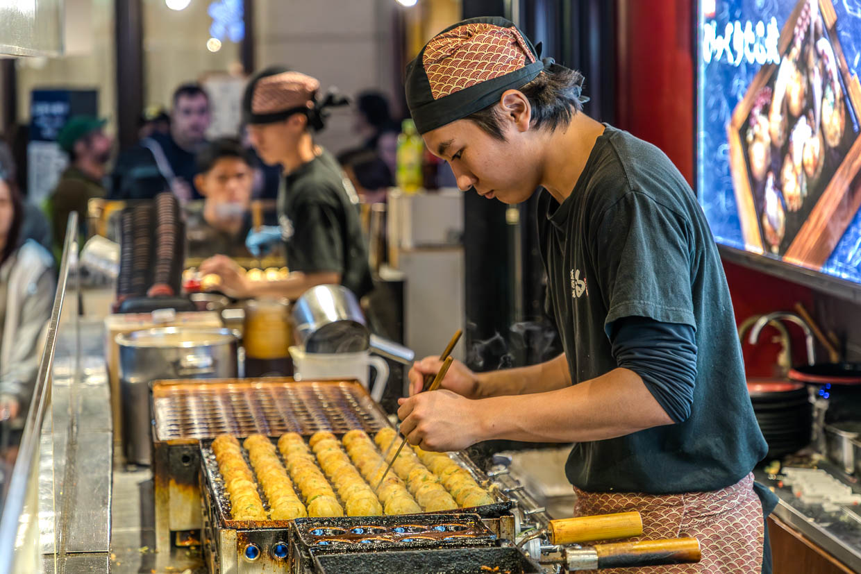 Koch bei der Zubereitung von Takoyaki. Kleine Oktopus-Stücke, grüne Zwiebeln und eingelegter Ingwer werden im Teigmantel in speziellen Backformen ausgebacken und vom Koch mit Stäbchen in großer Geschwindigkeit in der Form gewendet / © Foto: Georg Berg