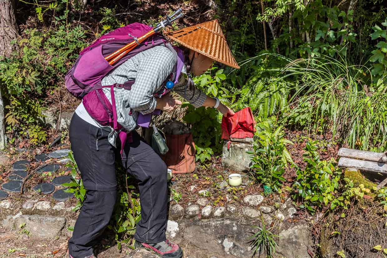 Guide zeigt einen Spalt unter dem roten Stoff des Jizo. Legt der Pilger eine Münze in diesem Spalt, so hilft das gegen Rückenschmerzen / © Foto: Georg Berg