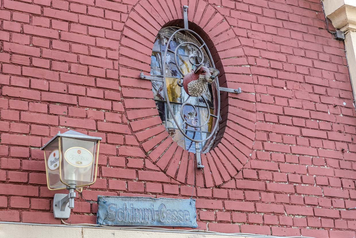 Provisorisches von Fans erstellte Straßenschild „Schimmi-Gasse“ hängt bis heute. Bevor die Horst-Schimanski-Gasse ein offizielles Straßenschild erhielt, gab es einen langjährigen Streit zwischen Fans und Stadtverwaltung. Die Gasse selbst (ursprünglich namenlos) wurde erst 2014 nach heftigen Streits benannt – gegen Widerstand der Stadtverwaltung wegen “fiktiver Figur” und Verwechslungsgefahr mit Fußballer Horst Szymaniak (“Schimmi”). Gestrickter Fuchs hängt am Fenster in der Schimmi-Gasse / © Foto: Georg Berg