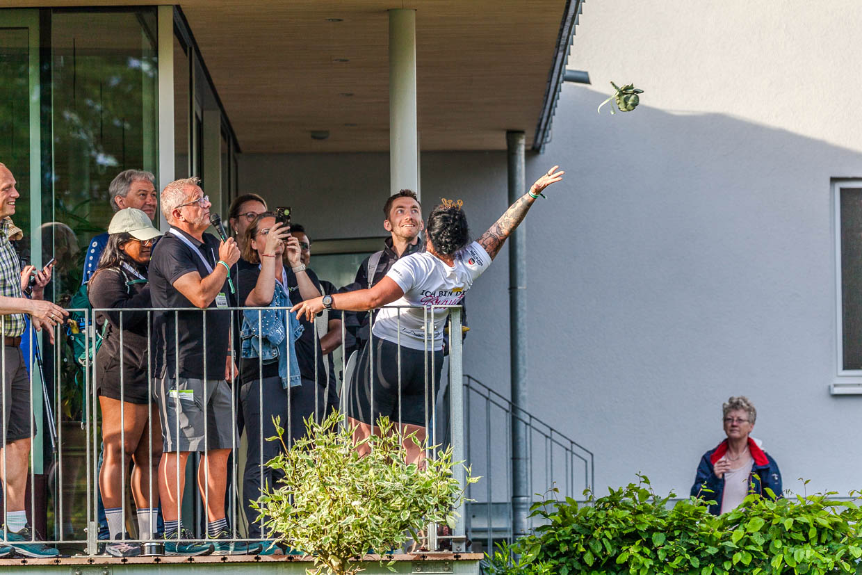 Eine Frau wirft ein kleines Bündel von einem Balkon, während eine Gruppe Menschen zuschaut in der Edwin-Trebes-Straße, Steinbach am Wald, Germany
