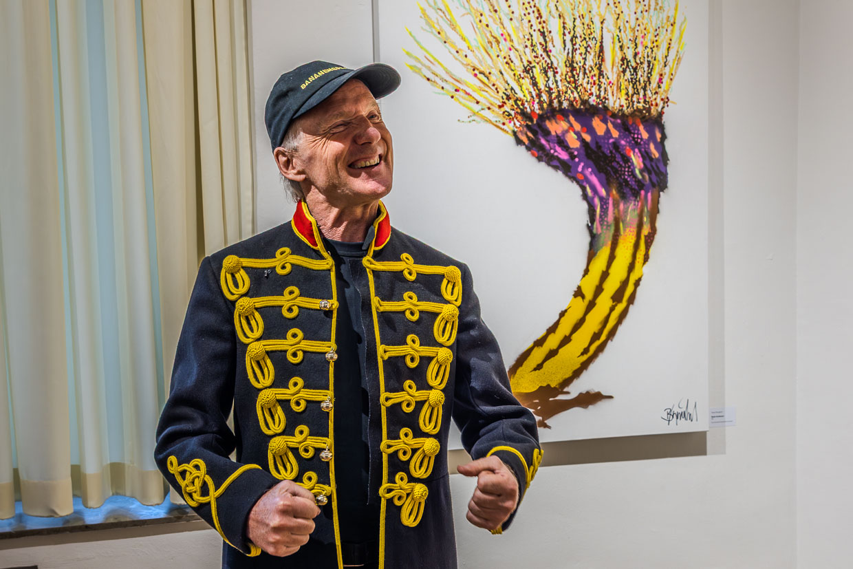 Ein lächelnder Mann in einer historischen Uniformjacke und Cap steht vor einem Kunstwerk auf der Schanze in Grevenbroich, Deutschland