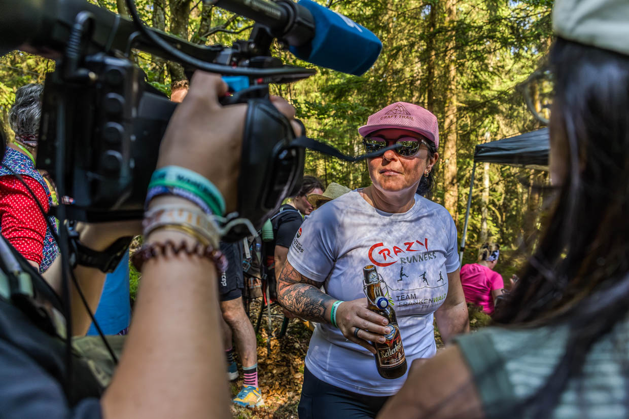 Frau mit CRAFT TRAIL Kappe und CRAZY RUNNERS Shirt hält Mönchshof Kellerbier in Steinbach am Wald während sie gefilmt wird