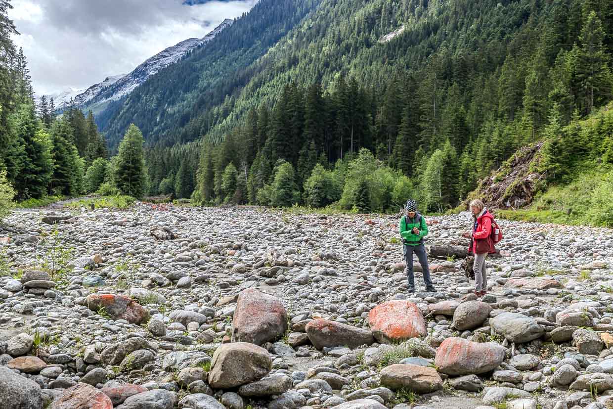 Zwei Wanderer erkunden das steinige Flussbett am Wasserfallrundweg Untersulzbach, Rosental, Austria, umgeben von bewaldeten Bergen und schneebedeckten Gipfeln